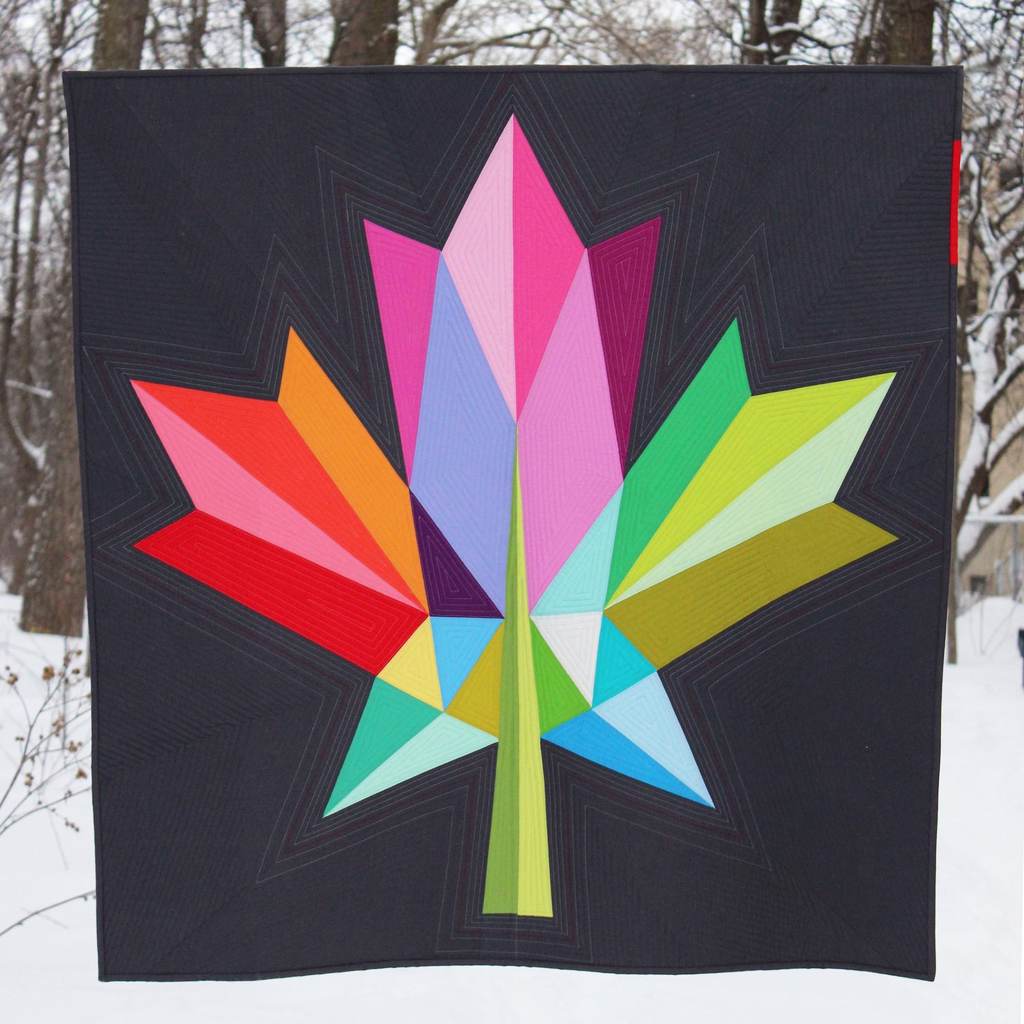 quilt of a modern bold maple leaf in multi colours on a dark grey ground held against a winter forest background