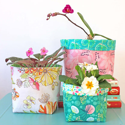 3 fabric baskets in colourful floral fabrics, housing flowering plants, stacked on books on n aqua surface