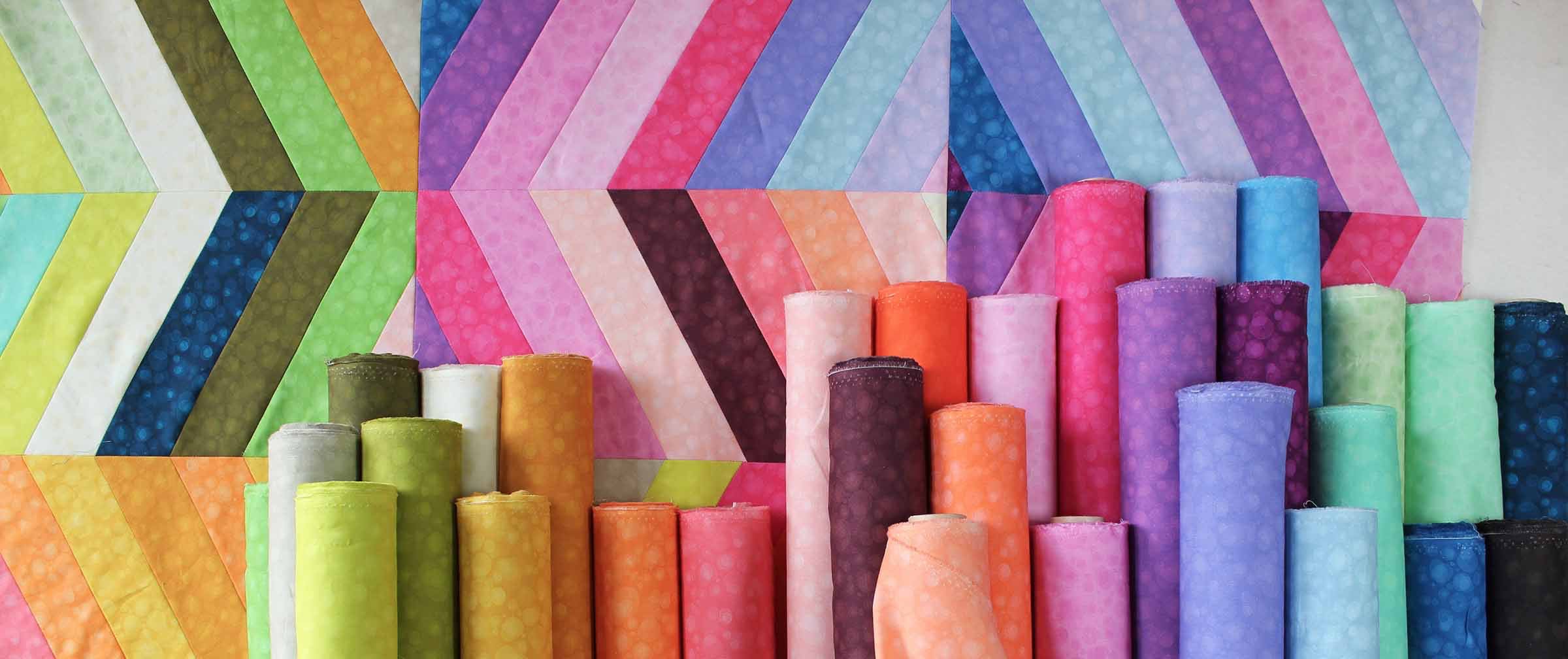 Colorful fabric rolls with a multi-coloured quilt on the wall in the background